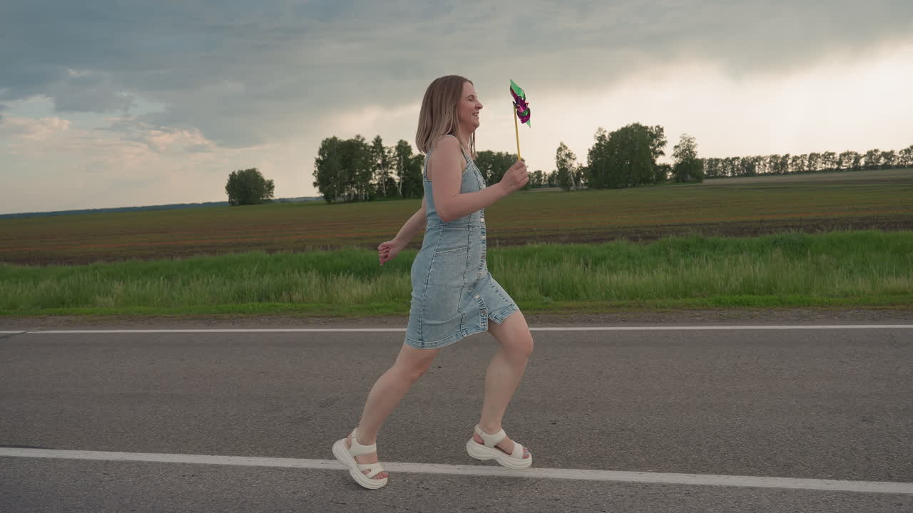 Side view of happy woman in denim dress running joyfully along straight asphalt road marked by white line holding colourful pinwheel high under cloudy summer sky hair blowing in gentle breeze