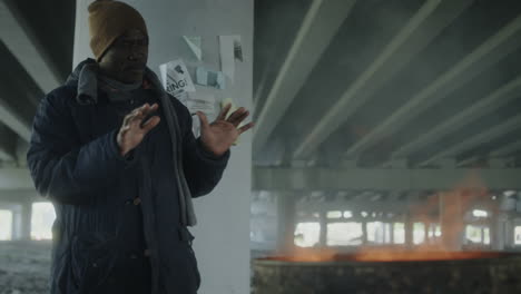 homeless man warming hands by fire under a bridge