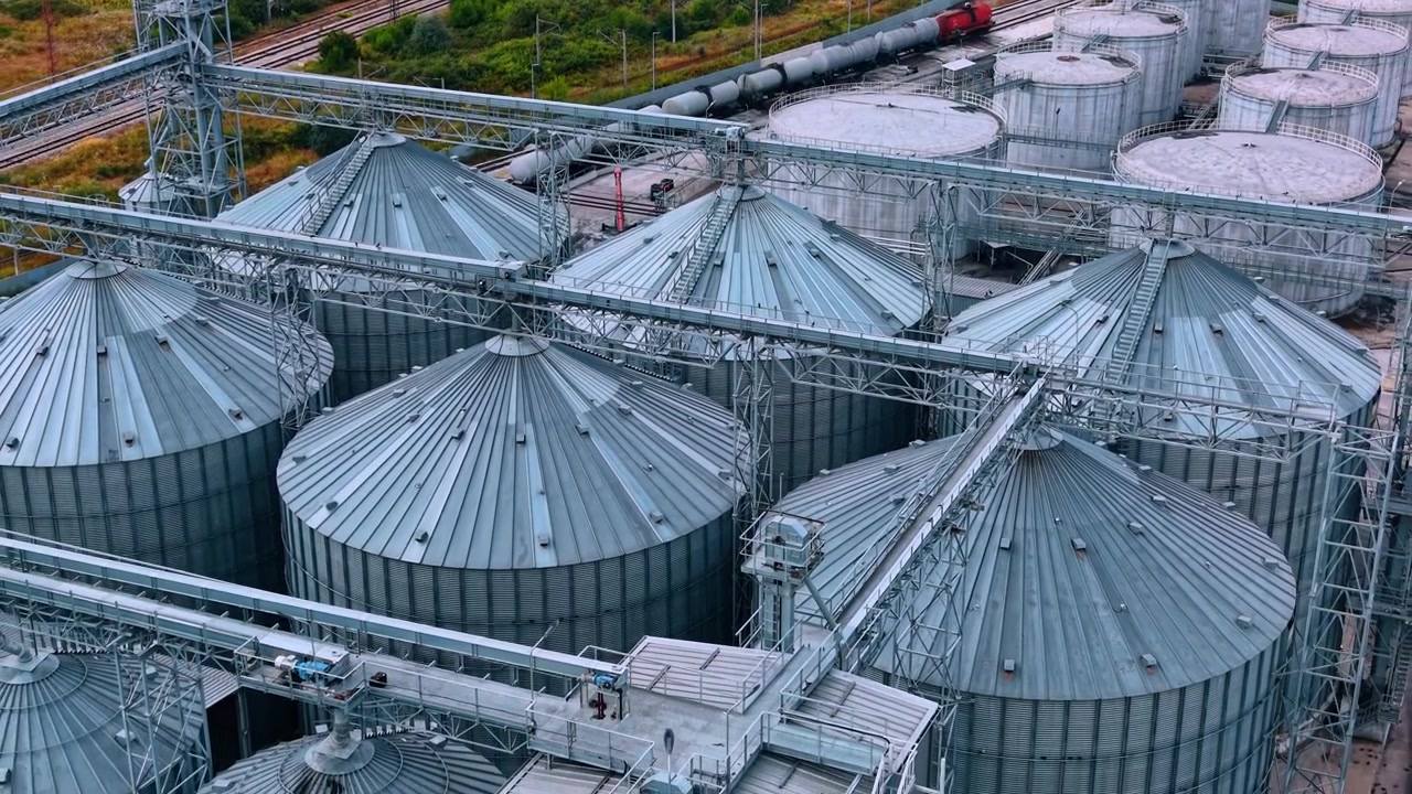 Storage tanks and rail tracks at a grain facility in a rural area. Large silver storage tanks are seen at a grain facility during a sunny day. Rail tracks run nearby.