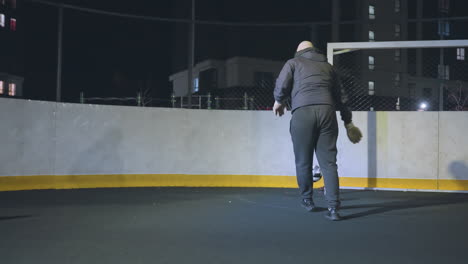 atleta en ropa deportiva visto desde atrás hábilmente pateando la pelota contra la pared cerca del poste de la portería en la cancha al aire libre por la noche con edificio residencial y telón de fondo urbano iluminado por luces brillantes