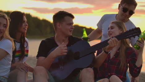 The-man-is-playing-the-guitar-on-the-open-air-party-with-his-friends-with-beer.-This-is-wonderful-evening-around-bonfire.