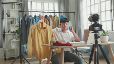 asian man online seller recommending a shirt and recording video by camera while using computer for selling clothes at home