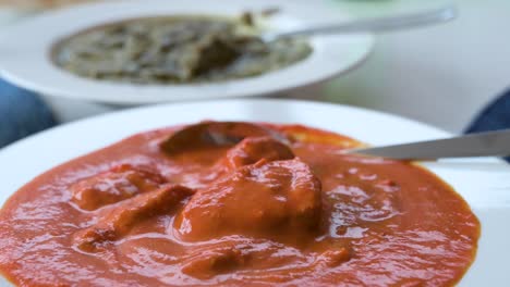 View-of-an-Indian-chicken-tikka-masala-dish,-consisting-of-roasted-marinated-chicken-chunks-in-a-spiced-orange-sauce,-seen-on-a-white-plate-at-the-forefront