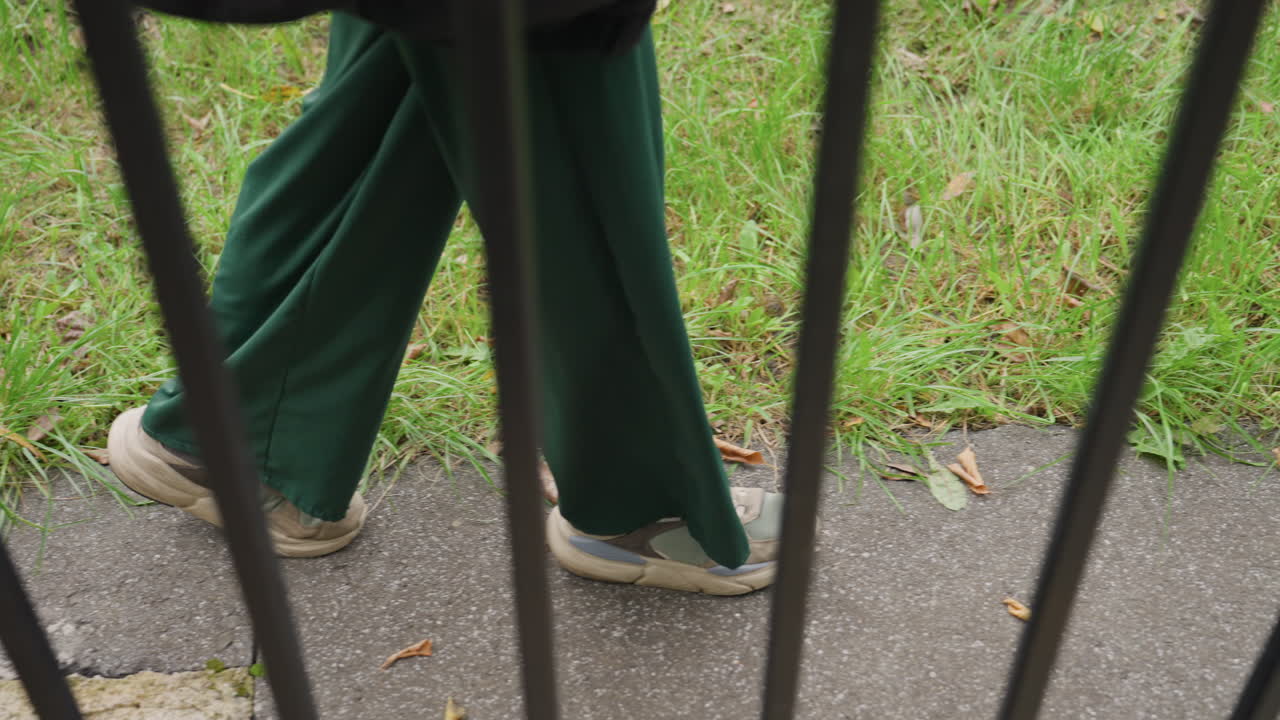 Pantaloni verdi, persona che cammina dietro una recinzione lungo un marciapiede crepato, scarpe da ginnastica usurate che sfregano sul selciato, foglie autunnali sparse sull'erba, barre di ferro verticali che creano una barriera incorniciata, inquadratura dal basso delle gambe