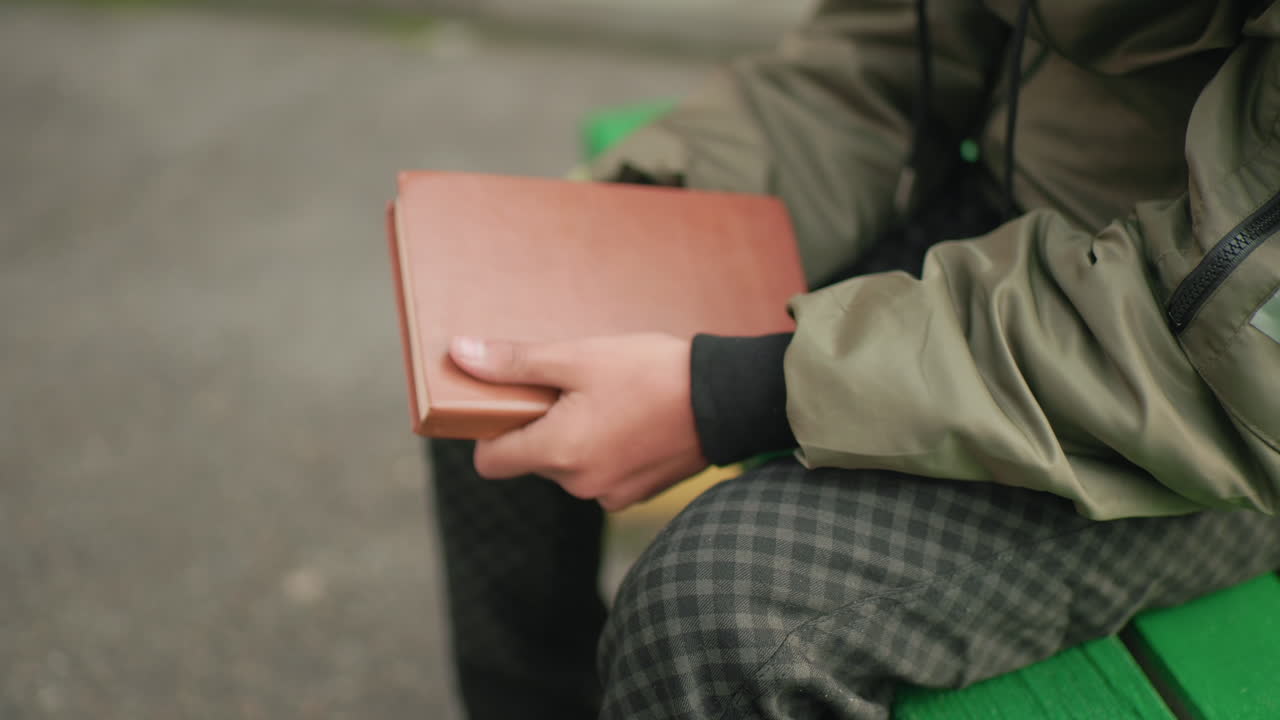 Close up side view of person in checkered pants and green jacket seated on bench holding brown cover novel in right hand gently tapping edge showing reflective action