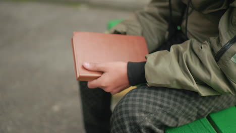 close up side view of person in checkered pants and green jacket seated on bench holding brown cover novel in right hand gently tapping edge showing reflective action