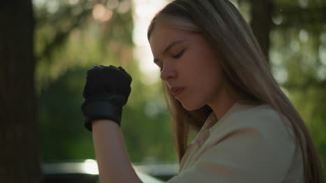 young adult wearing black gloves scratches elbow gently, sunlight filters through lush leaves creating a warm glow, blurred background features trees and greenery