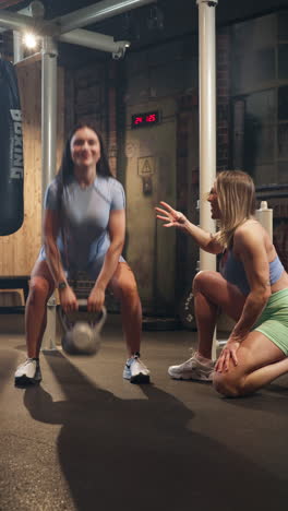 vertical, caucasian brown woman doing dumbbell deadlift while blond woman friend count the reps at the gym, indoor, handheld