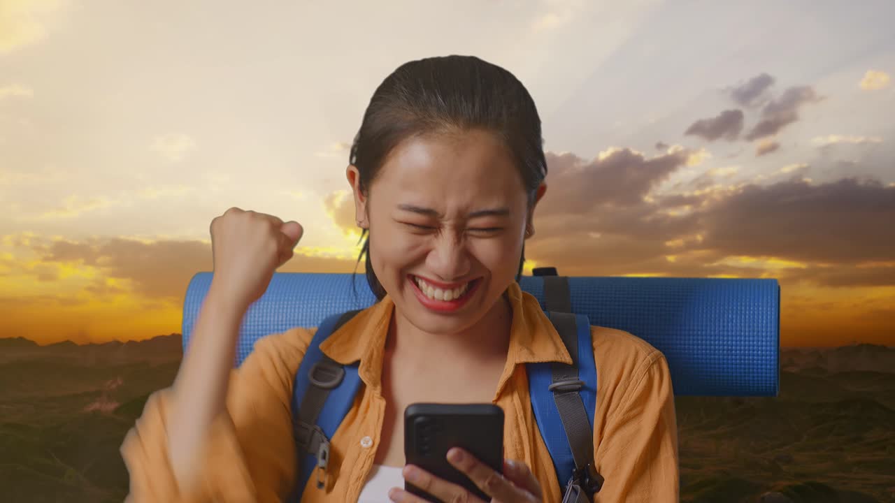 Close Up Of Asian Female Hiker With Mountaineering Backpack Looking At The Smartphone And Screaming Goal Celebrating The Success While Standing On The Top Of Mountain During Sunset Time