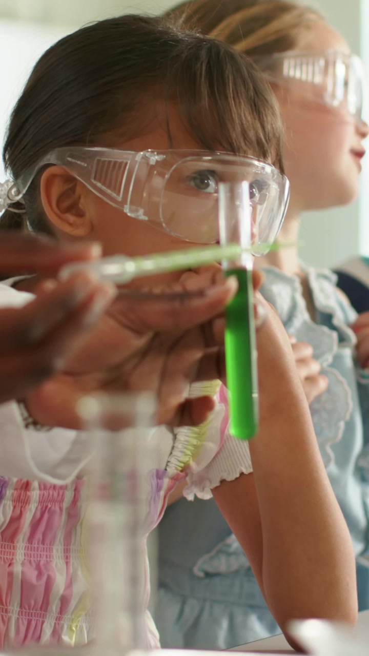 Vertical video: Leaning goggled girl in dress watching adult holding green tube at lab, copy space