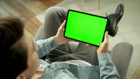 sobre el hombro vista joven en casa sentado en una silla usando con pantalla de maqueta verde tableta. hombre usando dispositivo de pantalla táctil, navegación por internet. concepto de pantalla verde y clave de croma