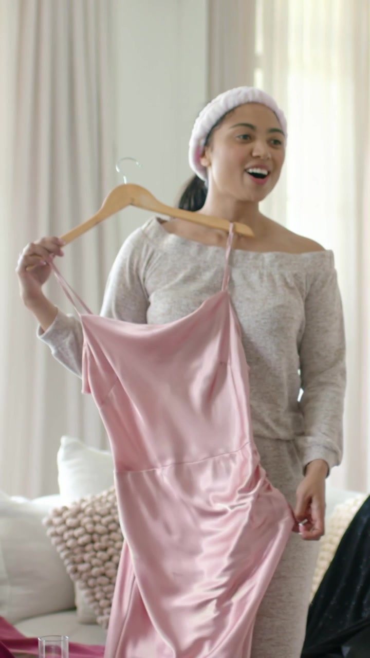 Vertical video: Taking woman holding pink satin dress on wood hanger from rack at home, gauging fit