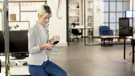 holding coffee mug, woman using smartphone in modern office setting