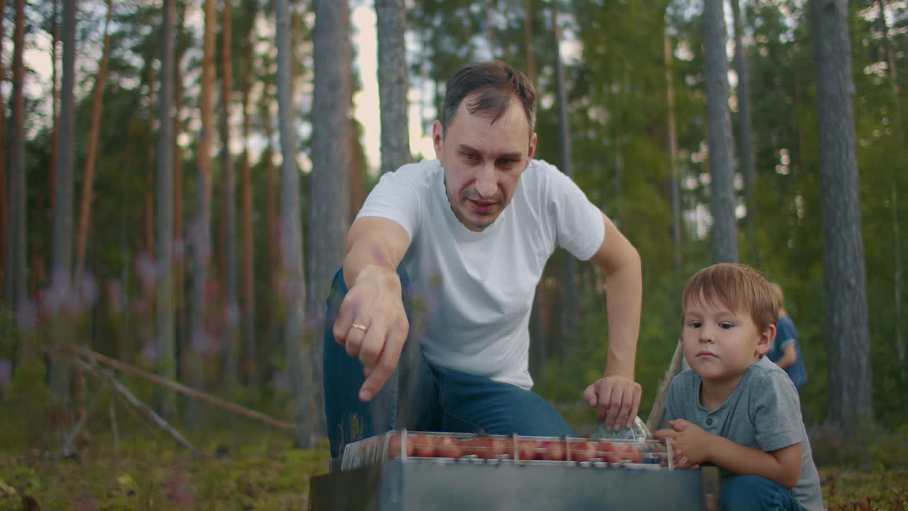 un ragazzino sta guardando come suo padre griglia salsicce. un fine settimana in natura. un uomo e un bambino si riposano nel bosco.