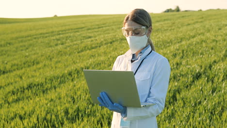 caucasian researcher woman in white coa, mask and goggles using laptop and looking around in the green field