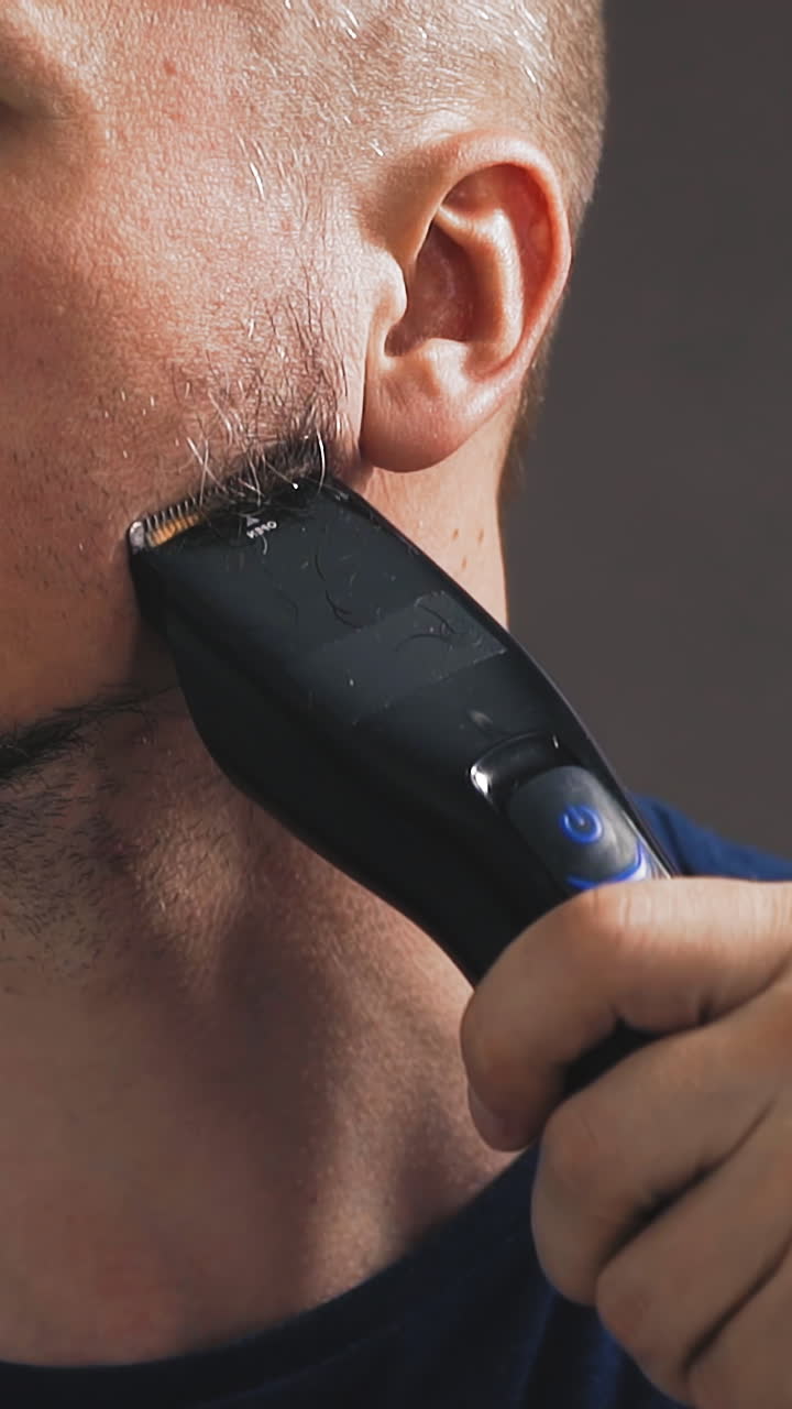 slow motion closeup portrait of young man shaves off beard grown during self-isolation and quarantine, looks at himself in mirror, holds hair cutter, in black footblock