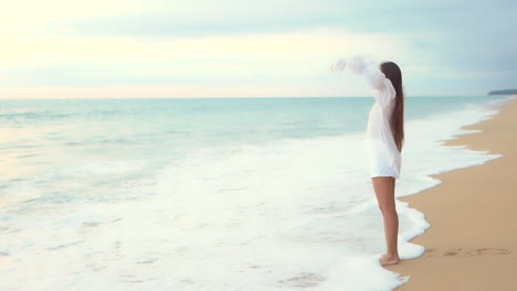 An-attractive-woman-in-a-white-coverup-walks-into-the-incoming-surf-raising-her-arms-in-joy