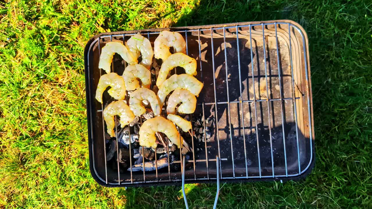 Close-up of a person's hand placing fresh raw shrimp on a portable charcoal grill on green grass