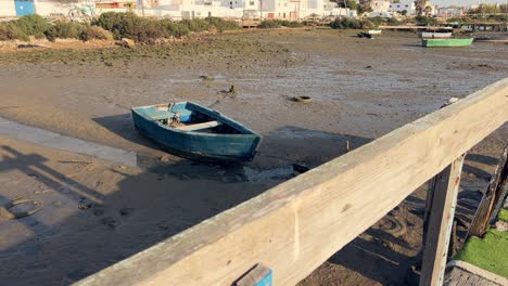 Moving-along-old-and-decayed-wooden-promenade-with-dried-up-and-muddied-ground-surface,-old-fishing-boat-and-tires-at-river-empty-bottom