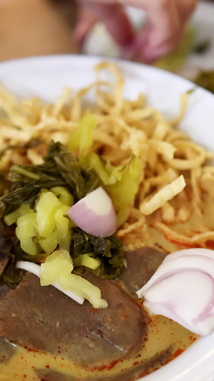 Una mano decora un tazón de fideos de curry de ternera del norte de Tailandia con chalotas en rodajas, vegetales encurtidos y fideos crujientes bajo una iluminación brillante y natural.