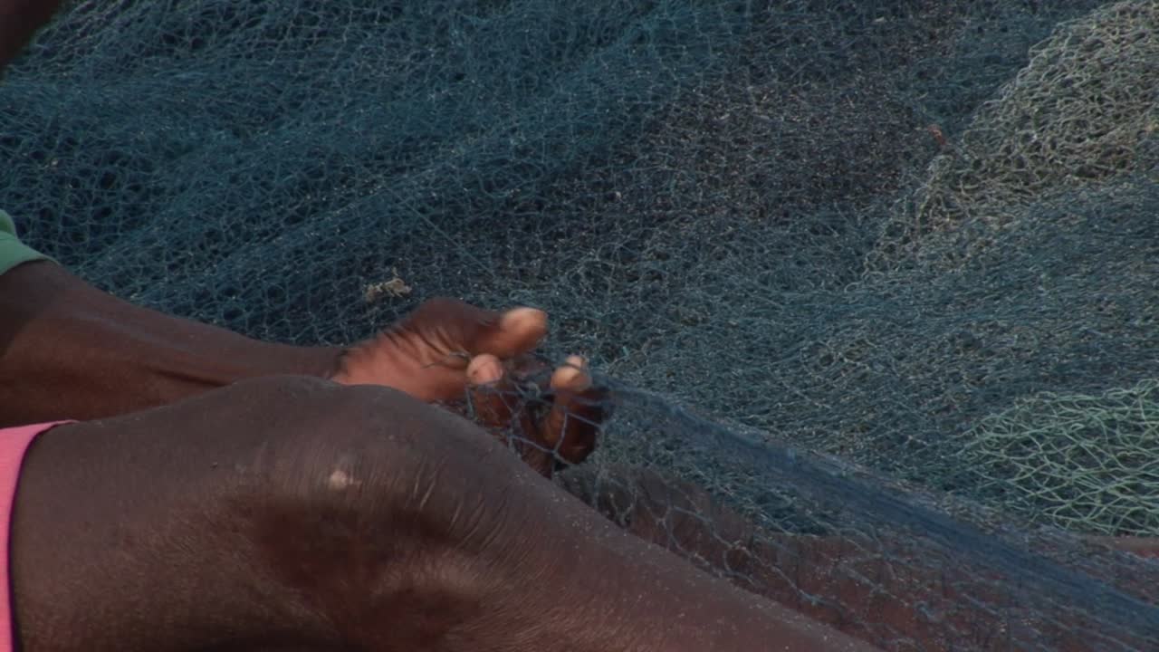 Primo piano delle mani di un pescatore ghanese che cuce e ripara rapidamente reti da pesca blu sulla spiaggia, evidenziando l'artigianato tradizionale, la texture e il sostentamento costiero