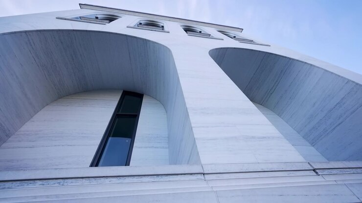 Uma estrutura arquitetônica moderna com fachada de concreto e vidro, capturada de um ângulo baixo, olhando para o design limpo e brilhante do edifício em um ambiente urbano com céu claro.