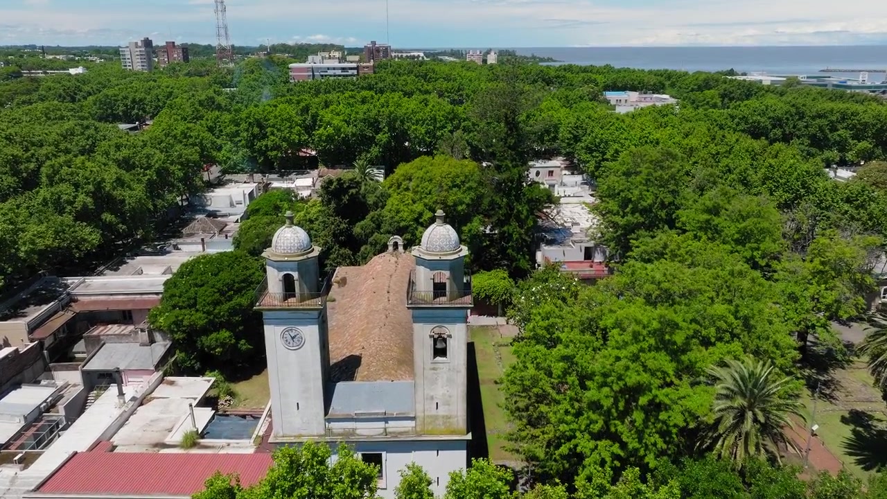 Basilica of the Holy Sacrament in Colonia del Sacramento Uruguay, drone establishing dolly at midday with vibrant lush green trees and white low buildings