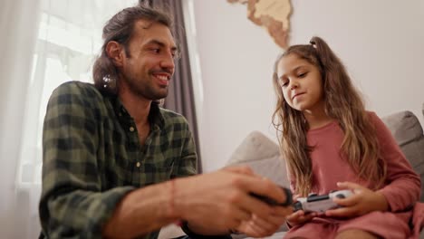 Close-up-shot-of-a-brunette-man-in-a-green-checkered-shirt-playing-and-telling-how-to-properly-hold-a-joystick-in-his-hands-together-with-his-daughter,-a-little-brunette-girl-in-a-pink-dress-on-a-gray-sofa-in-a-modern-apartment