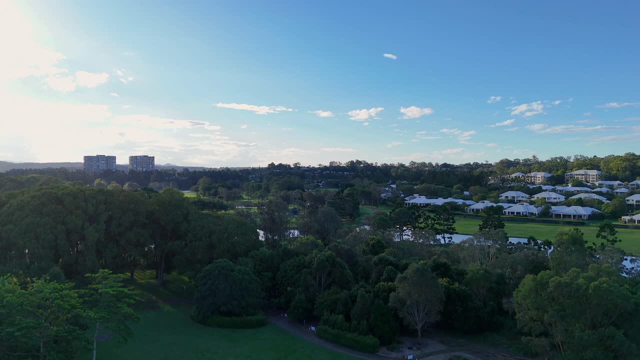 Drone footage captures a lush golf course with surrounding greenery and distant residential areas under a clear sky