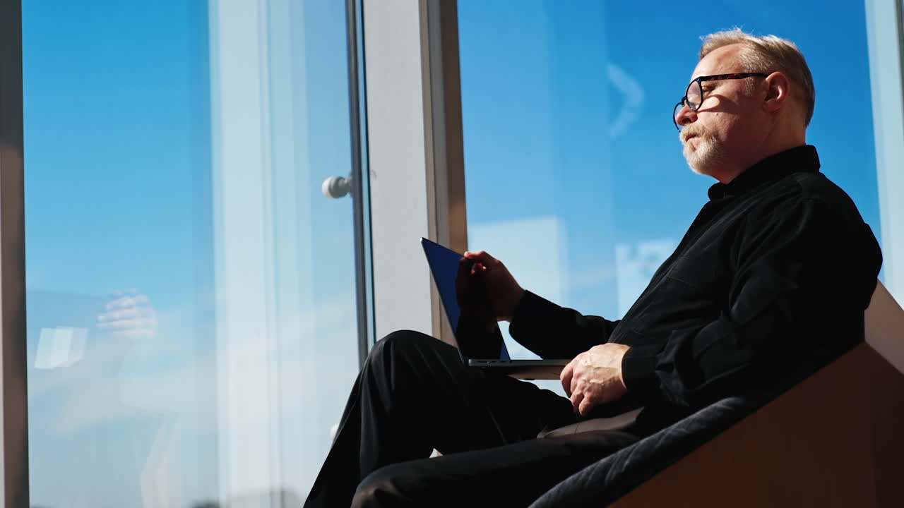 Pensive Caucasian man wearing black clothes and eyeglasses sits in the armchair. Businessman works on laptop and looks at window. Low angle view.