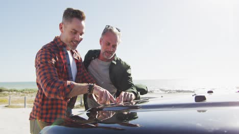 Happy-caucasian-gay-male-couple-leaning-on-car,-reading-map-and-talking-on-sunny-day-at-the-beach