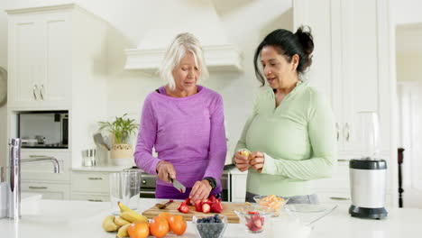 two happy diverse senior women chopping fruits and laughing in kitchen, slow motion