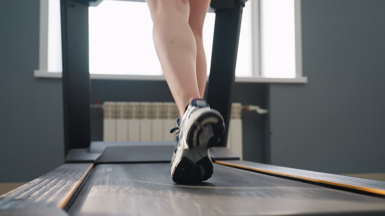 rear view of woman walking on treadmill in gym environment with one foot mid-air showing athletic movement on workout machine positioned near large windows and tiled floor