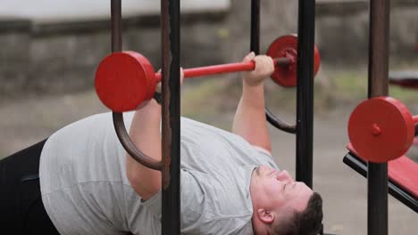 a persistent fat man lifts weights lying down on outdoor exercise machines a persistent fat man lifts weights lying down on outdoor exercise machines struggle with excess weight training man on the background of asphalt
