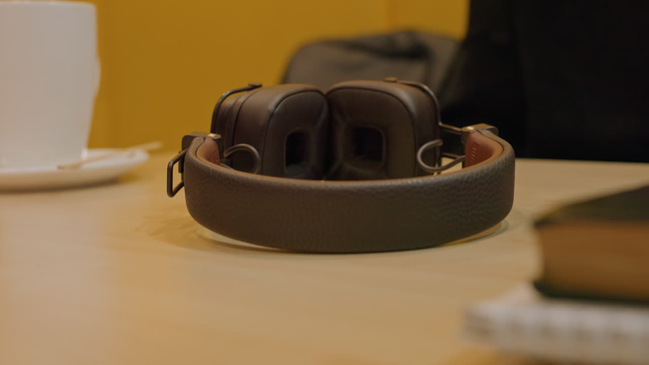 Headphone di meja kayu dengan orang kulit hitam di latar belakang, dinding kuning hangat, cangkir kopi dan buku catatan di dekatnya, headphone kulit terlipat, istirahat kreatif yang tenang, cahaya ambient lembut, ruang kerja intim