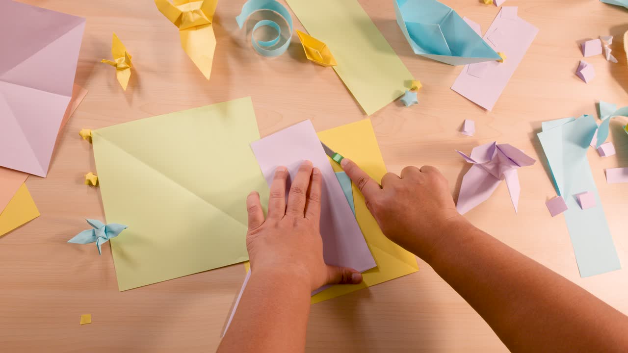 Person folds pastel paper for origami on wooden desk, surrounded by finished paper models