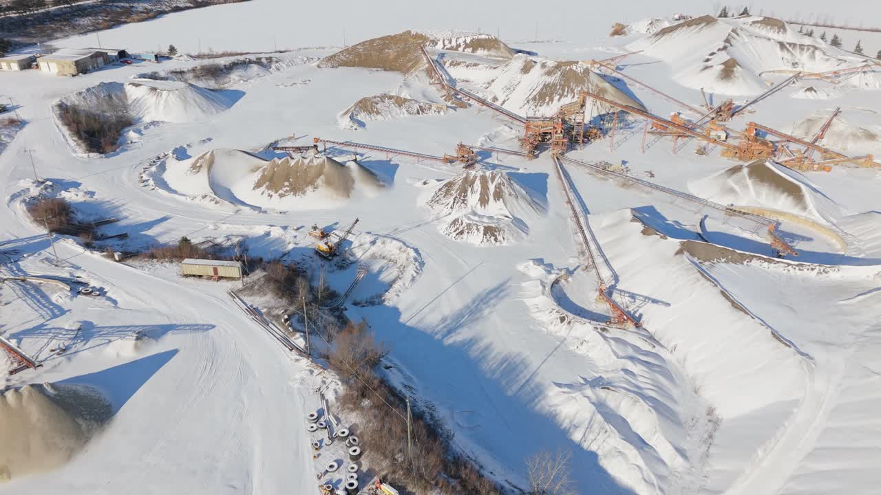 Gravel quarry with snow-covered mounds, conveyor belts, and machinery under winter skies