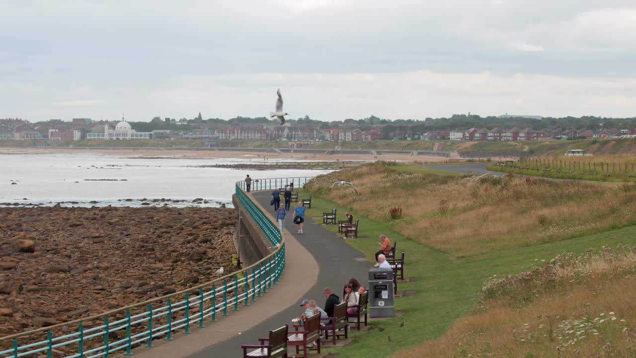 Several people stroll and sit along a curved seaside path bordered by grassy dunes, benches, and railings under overcast daylight, with distant town and shoreline visible