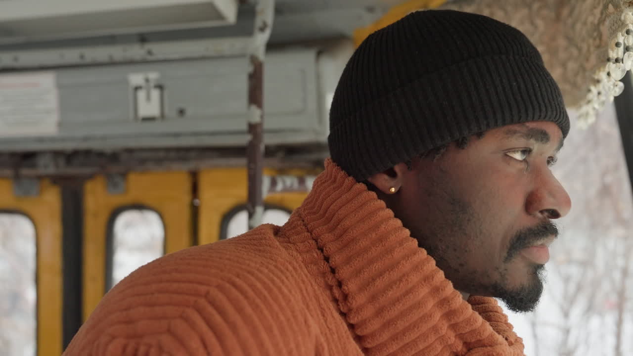 Hombre afroamericano mirando a través de una ventana escarchada, con suéter naranja y gorro negro, interior de autobús vintage, paisaje nevado exterior, luz natural suave, ambiente tranquilo y reflexivo, retrato de perfil en primer plano.