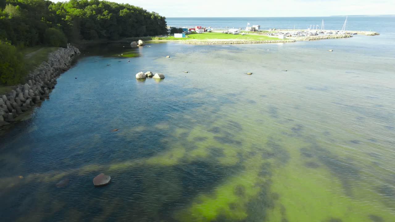 Aerial drone flyover footage of Tilgu seaside shoreline during a sunny summer day with a harbor with sailboats and yachts visible in the horizon besides grassy green shores that have rocky piers on it