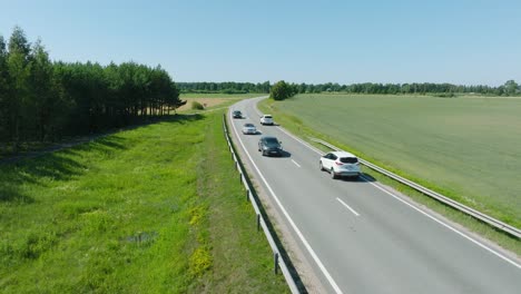 Aerial-establishing-shot-of-a-rural-landscape,-countryside-road-with-trucks-and-cars-moving,-lush-green-agricultural-crop-fields,-sunny-summer-day,-wide-ascending-drone-shot-moving-forward