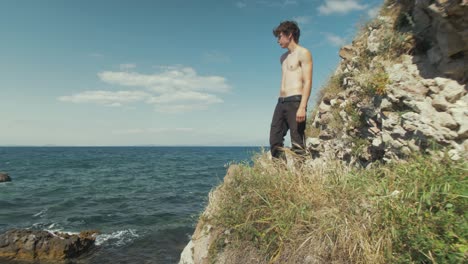 un joven caucásico sin camisa se para en un acantilado con vista al océano