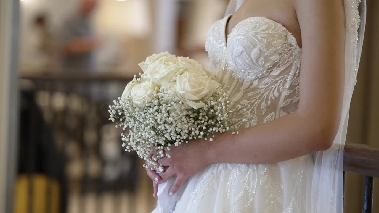 A bride stands holding a bouquet of white roses and baby's breath. She is dressed in a wedding gown. People can be seen in the background preparing for the ceremony