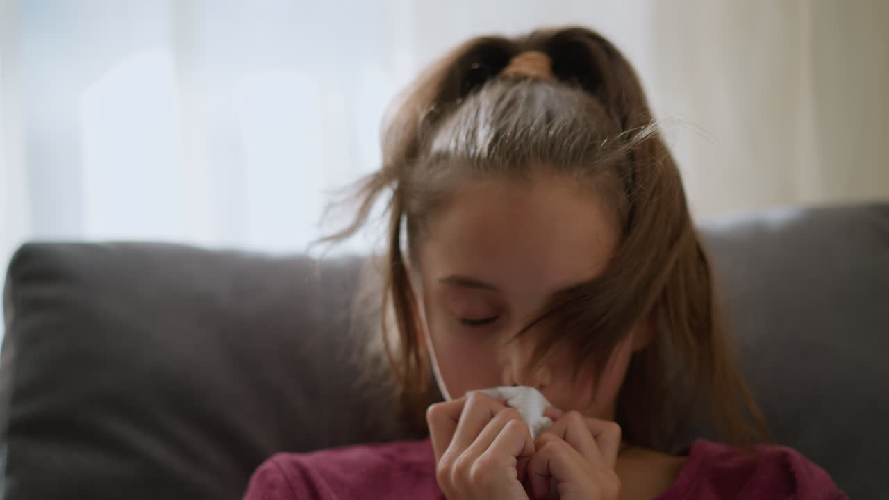 Close-up of little child with red bangle and tied hair sneezing into tissue due to flu, child appears unwell while lying on couch, showing signs of flu symptoms with tissue in hand