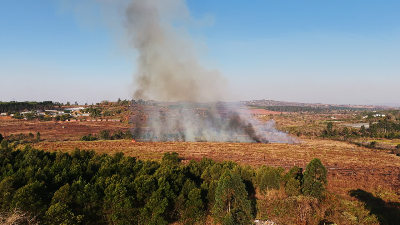 Aerial view of a grassland fire sending thick smoke into the sky, with surrounding fields and scattered buildings visible in the distance