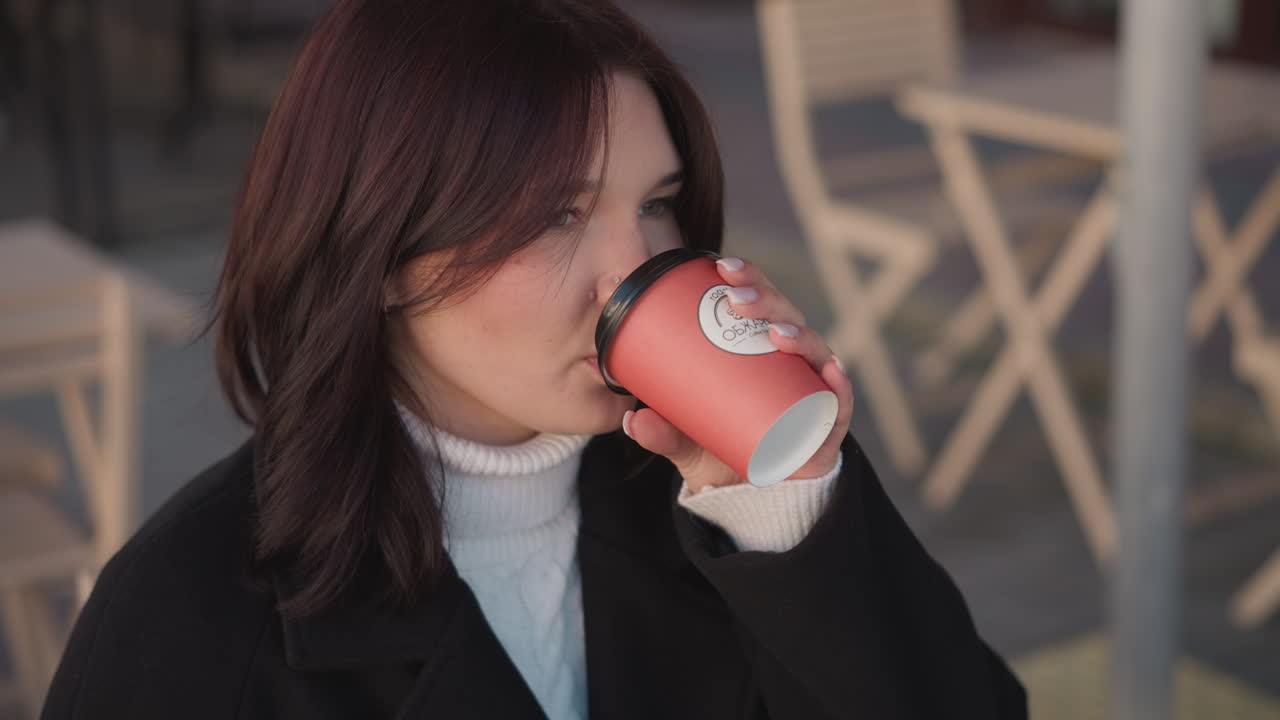 프리랜서가 커피를 마시며 컵을 테이블 위에 다시 부드럽게 놓고 나무 의자와 꽃의 흐릿함을 배경으로 쾌적한 야외 공간에서 일에서 휴식을 취합니다.