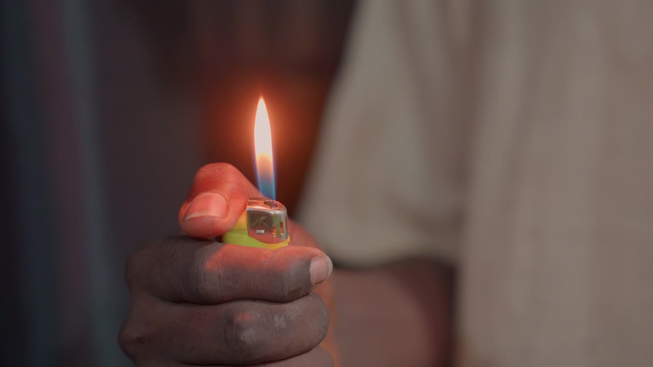 Soft closeup black hand holding lighter, gentle thumb lifts flint and blueorange flame warms beige shirt, evening indoor mood, careful gesture and tactile skin texture frame roles include camper