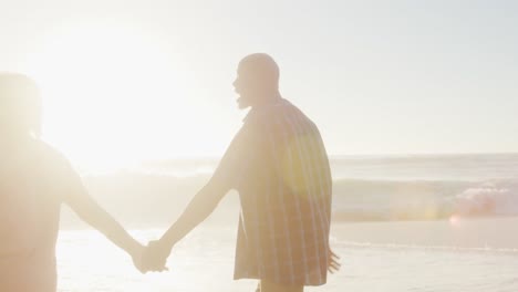smiling senior african american couple holding hands and walking on sunny beach
