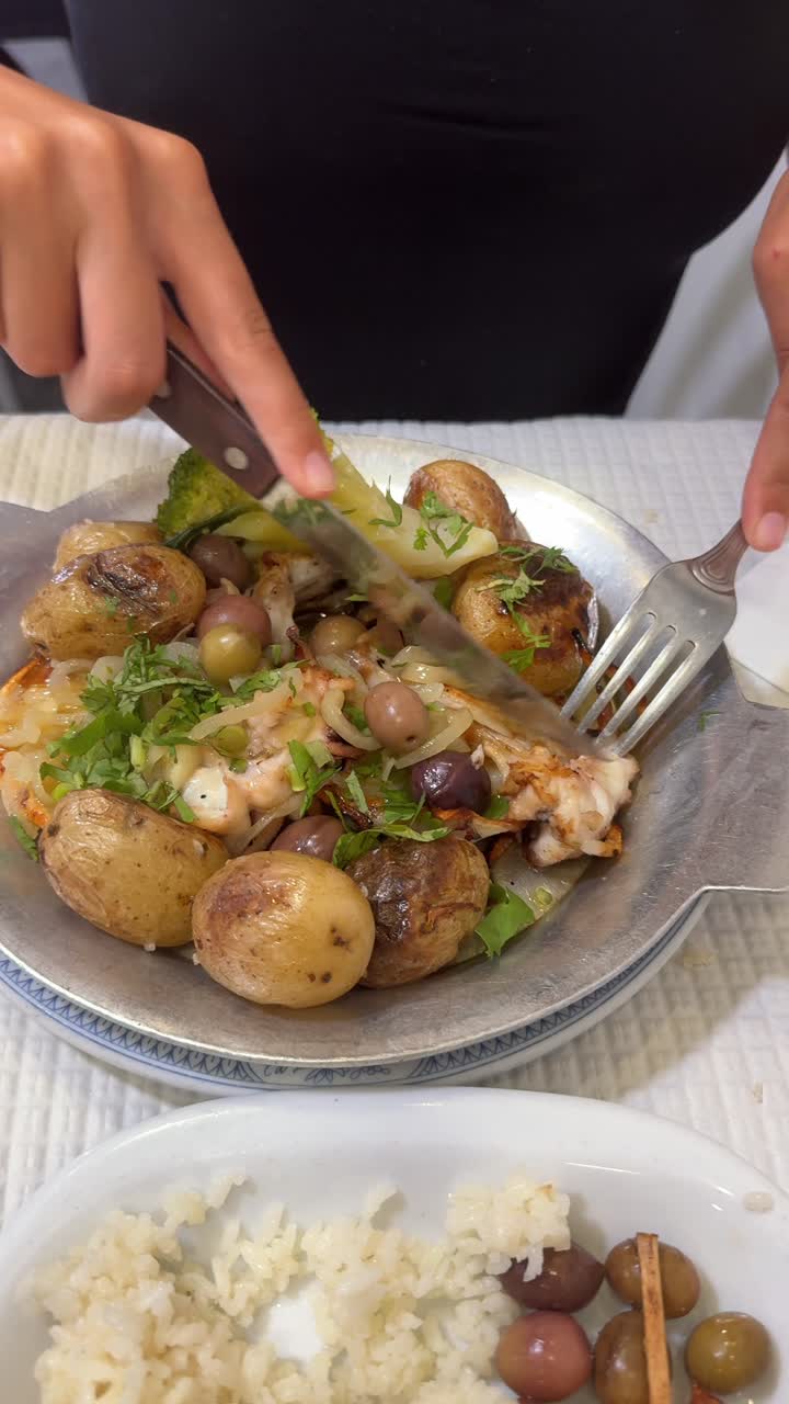 Person cutting a meal of roasted cod, potatoes, olives, and broccoli