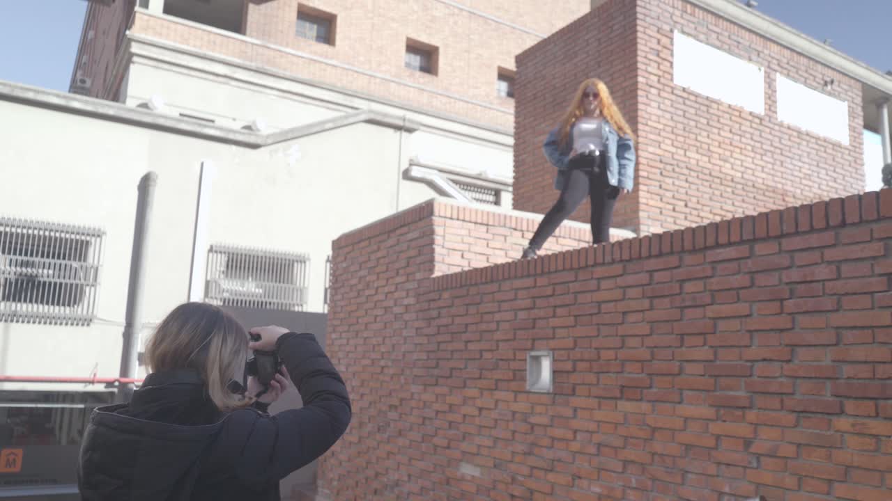 Two friends have fun taking pictures in the street. Redhead model poses on top of a brick wall and smiles for her photographer friend